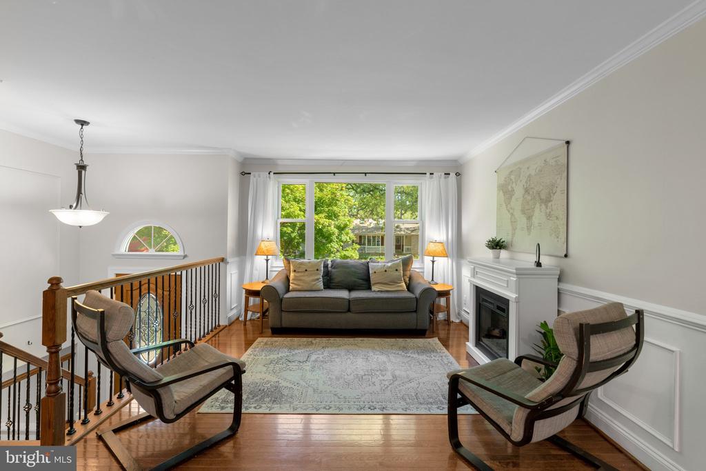 Upstairs living room with a gray sofa, white fireplace, hardwood floors, and large windows overlooking trees.