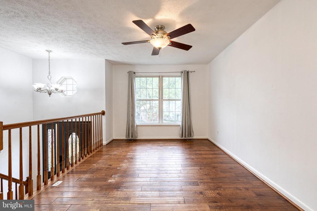 Empty upper-level room with dark hardwood floors, a curtained window, ceiling fan, and wooden stair railing.