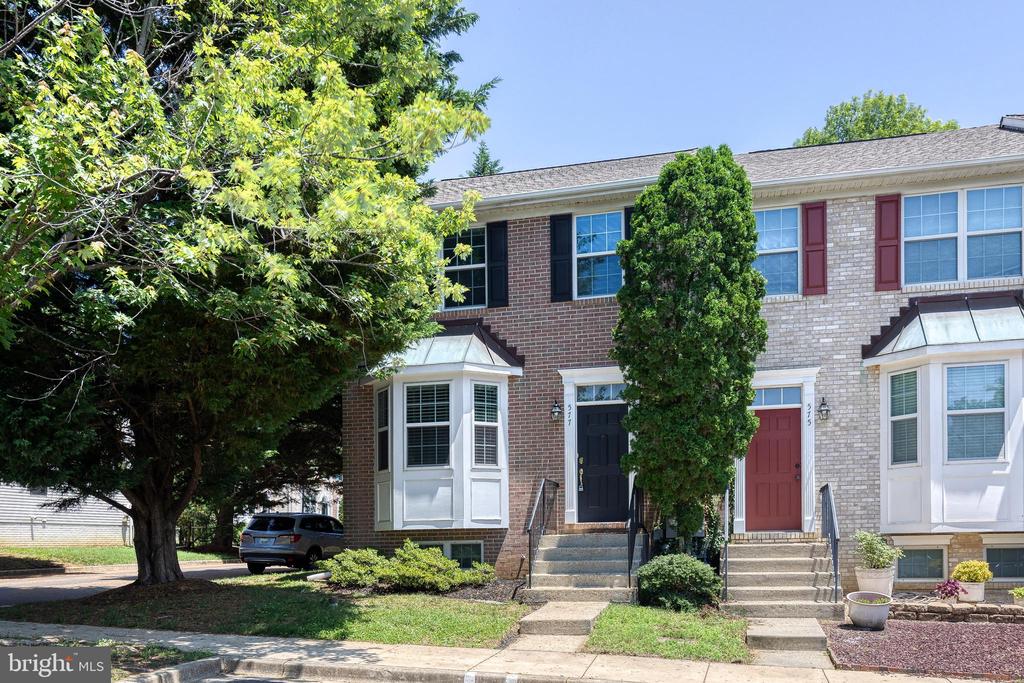 Brick townhouse with a projecting bay window, dark entry door, concrete steps, and mature shade trees.