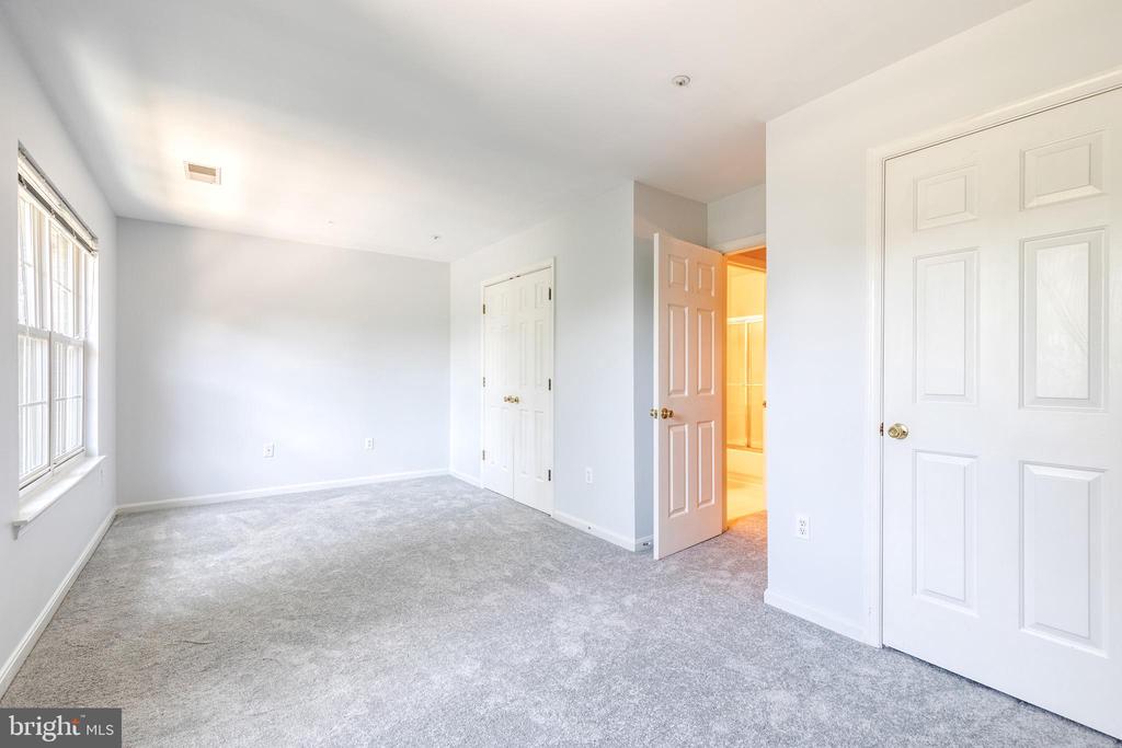 Empty bedroom with gray carpet, tall windows, and an open door to a brightly lit adjoining room.