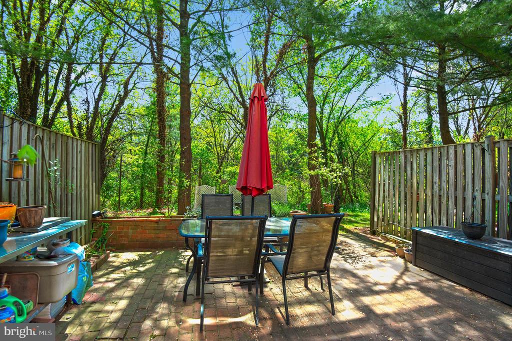 Fenced patio with a glass dining table, black chairs, and a closed red umbrella beneath leafy trees.