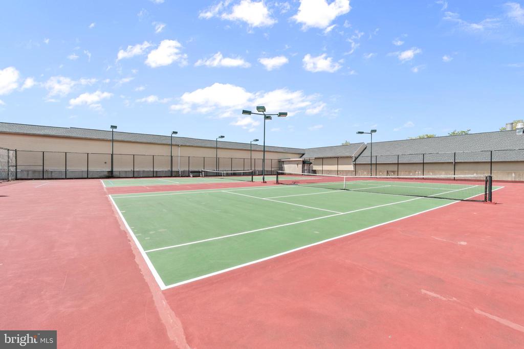 Outdoor tennis courts with green playing surfaces, red surrounds, and surrounding fencing.