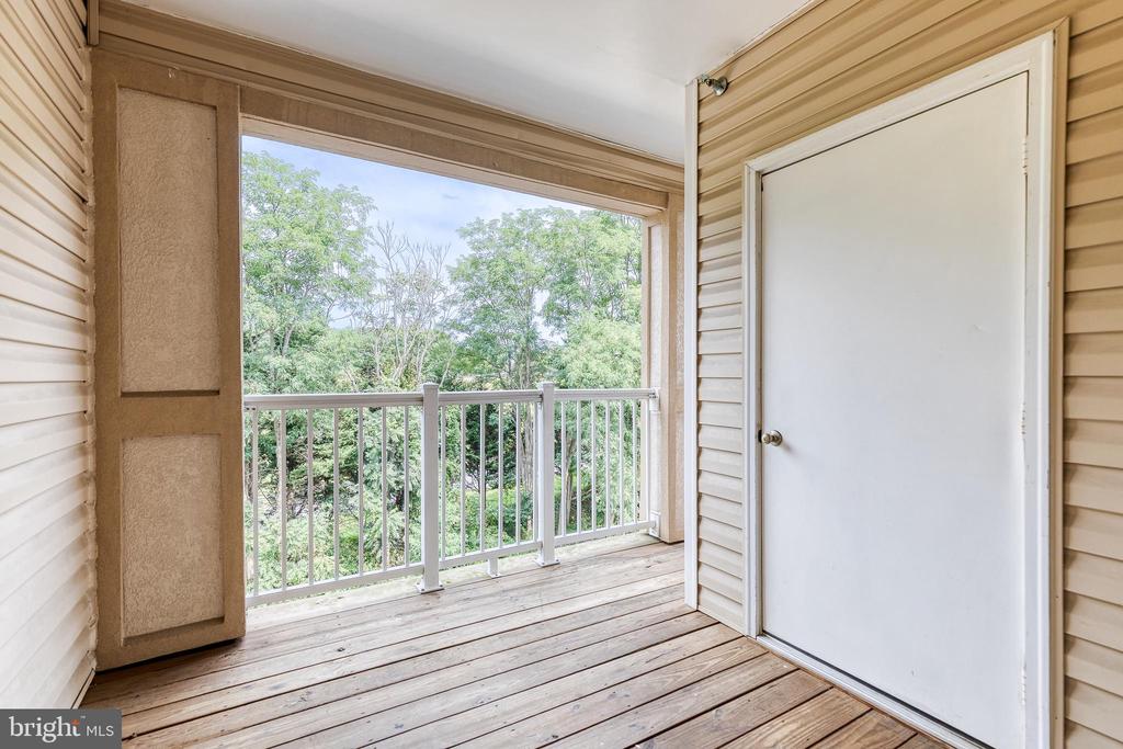Covered balcony with wood-plank flooring and white railing overlooking leafy trees.
