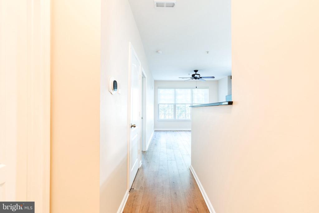 Vacant condo hallway with wood floors leading to a sunlit living area and kitchen counter.