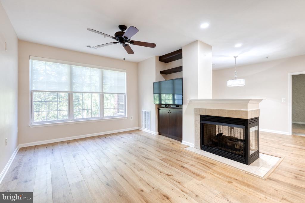 Empty living room with pale wood floors, a large window, built-in TV shelving, and a two-sided fireplace.