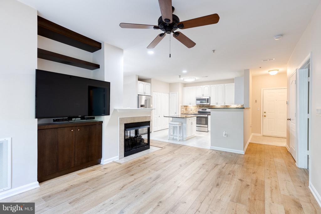 Open living room with a fireplace, wall-mounted TV, light wood floors, and adjoining white kitchen.