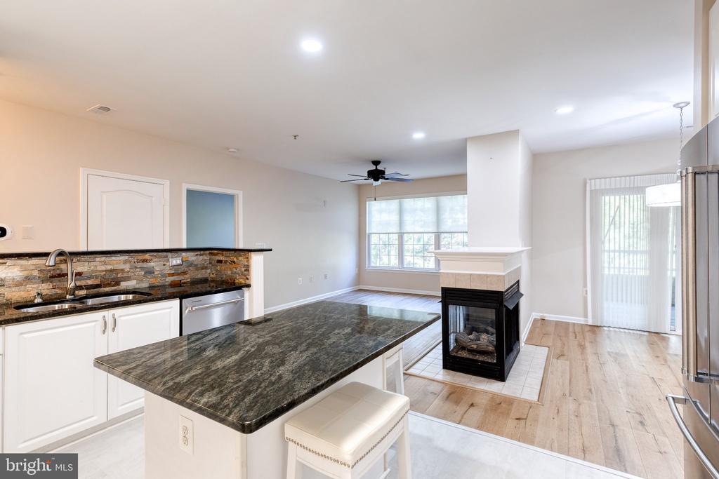 Open-plan kitchen with a granite island and stone fireplace beside an unfurnished living area.