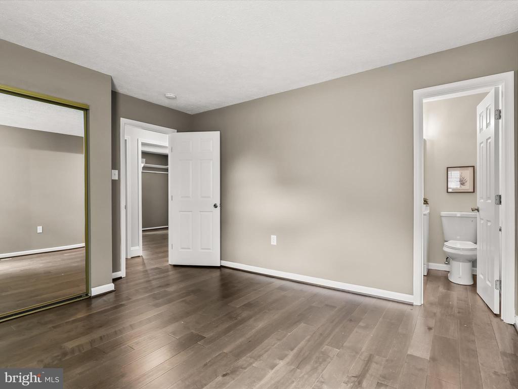 Empty bedroom with dark wood flooring, mirrored closet doors, and an adjoining bathroom.