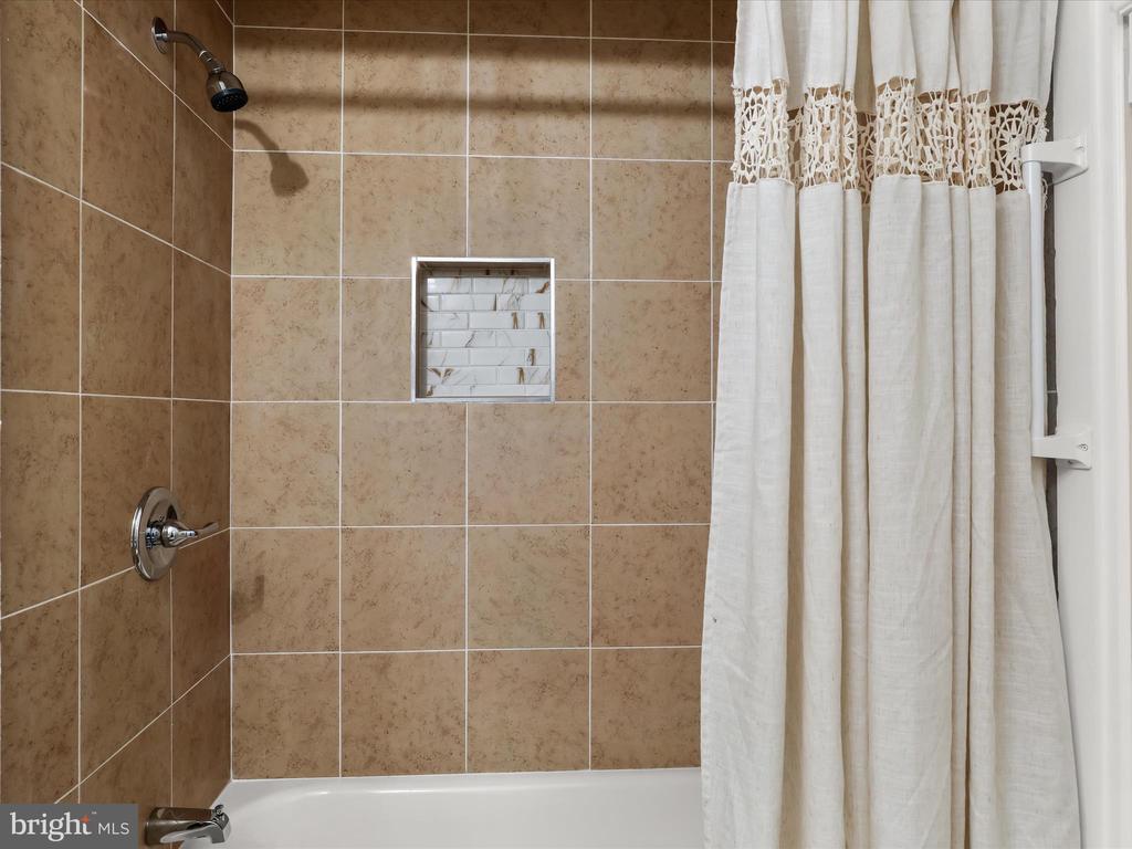 Bathroom with tan tiled shower surround, recessed niche, and white curtain over the bathtub.