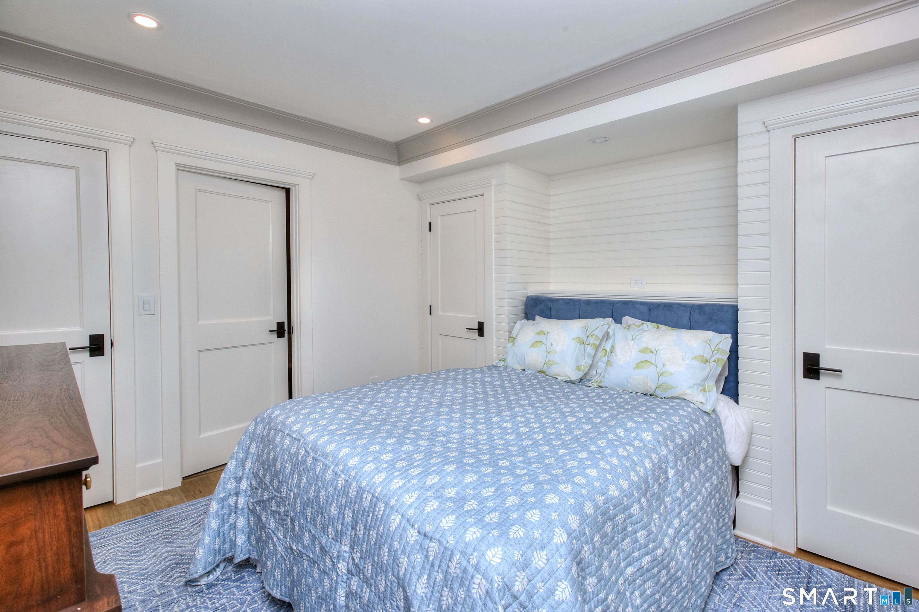 Bedroom with a blue quilted bed, upholstered headboard, and white paneled walls.