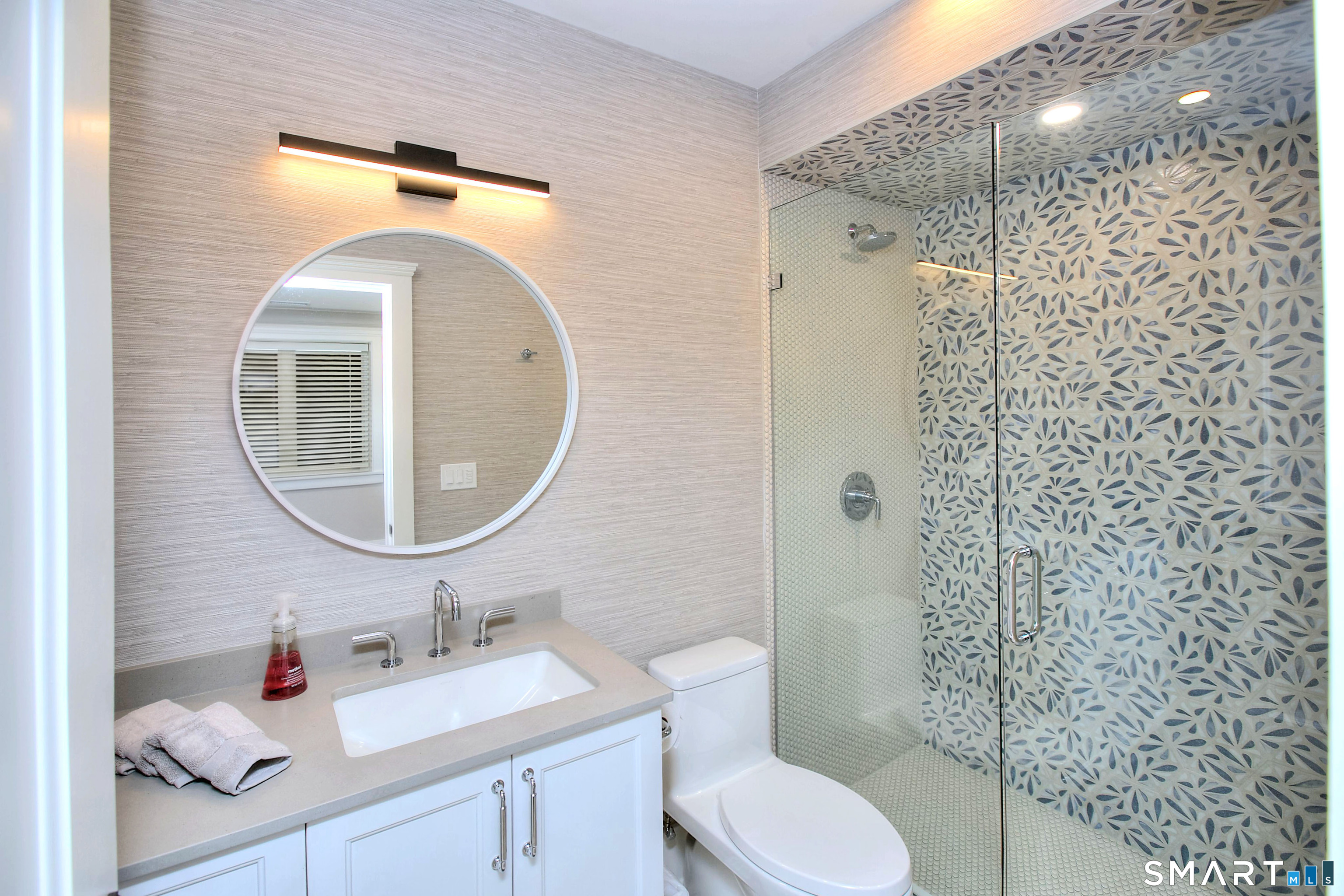 Modern bathroom with a round mirror, white vanity, and glass shower lined with blue-gray patterned tile.