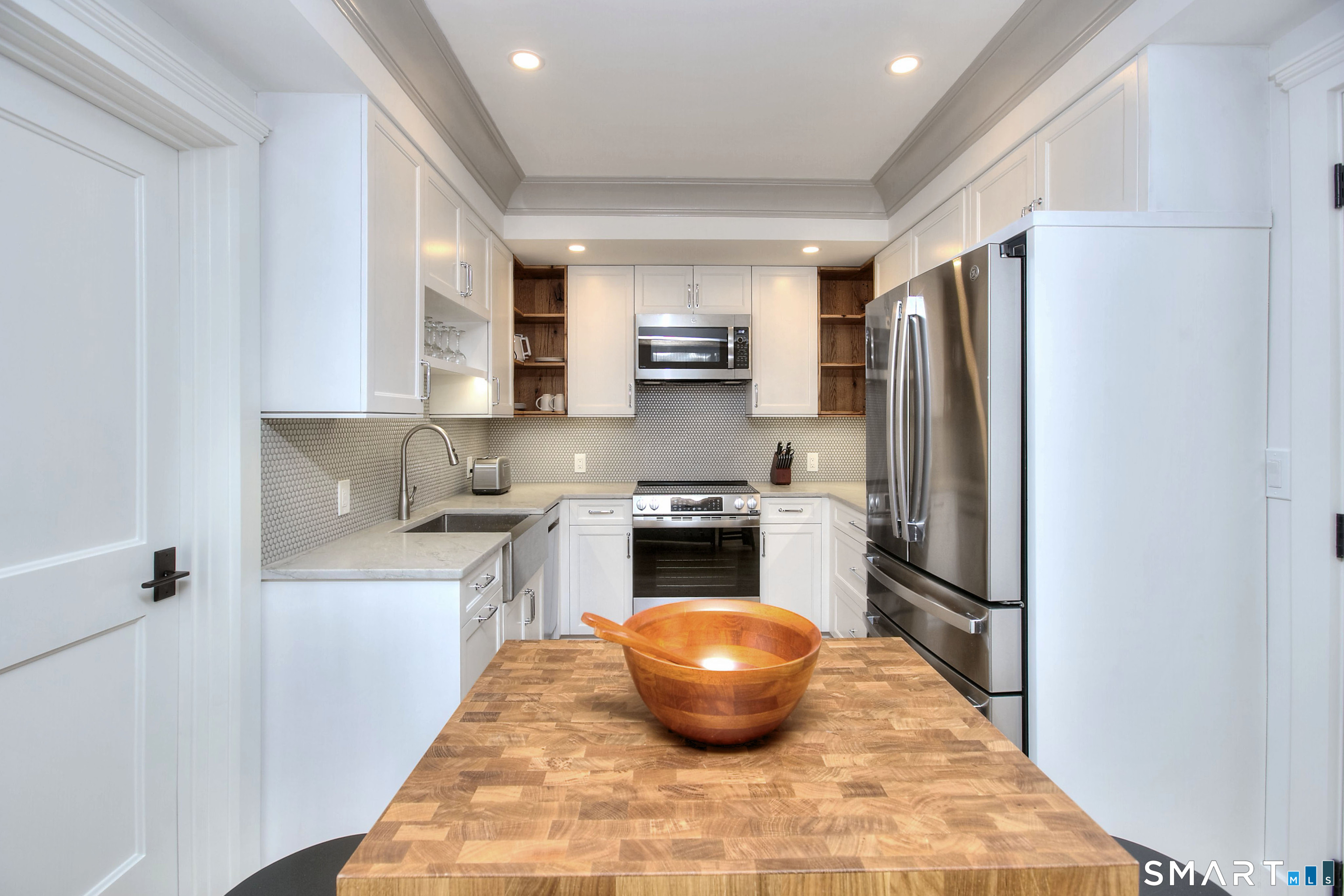 Modern white kitchen with stainless steel appliances and a butcher-block island topped by a wooden bowl.