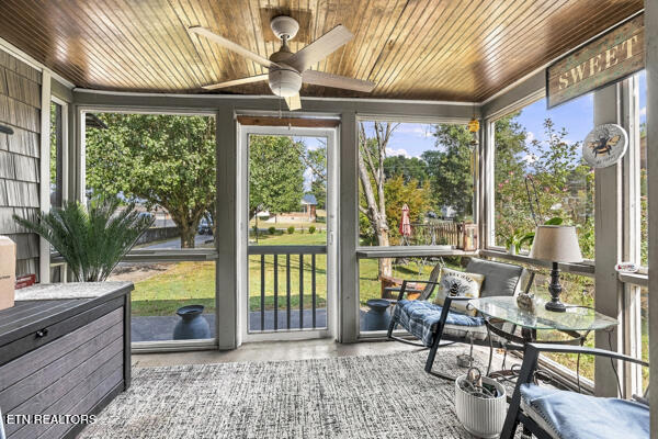 Enclosed porch with a wood ceiling, ceiling fan, and glass table overlooking a leafy lawn.
