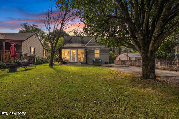 House at dusk with warmly lit windows, a broad grassy yard, and mature trees.