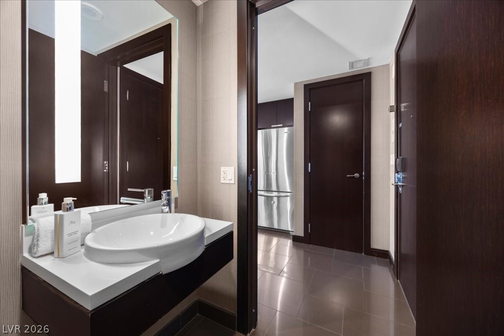 Bathroom vanity with oval vessel sink and mirror, opening onto a tiled hall with a stainless-steel refrigerator.