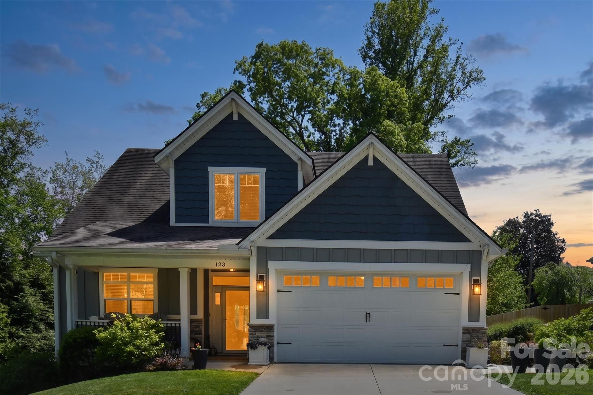 Two-story blue-gray house with a covered porch, glowing windows, and an attached garage at dusk.