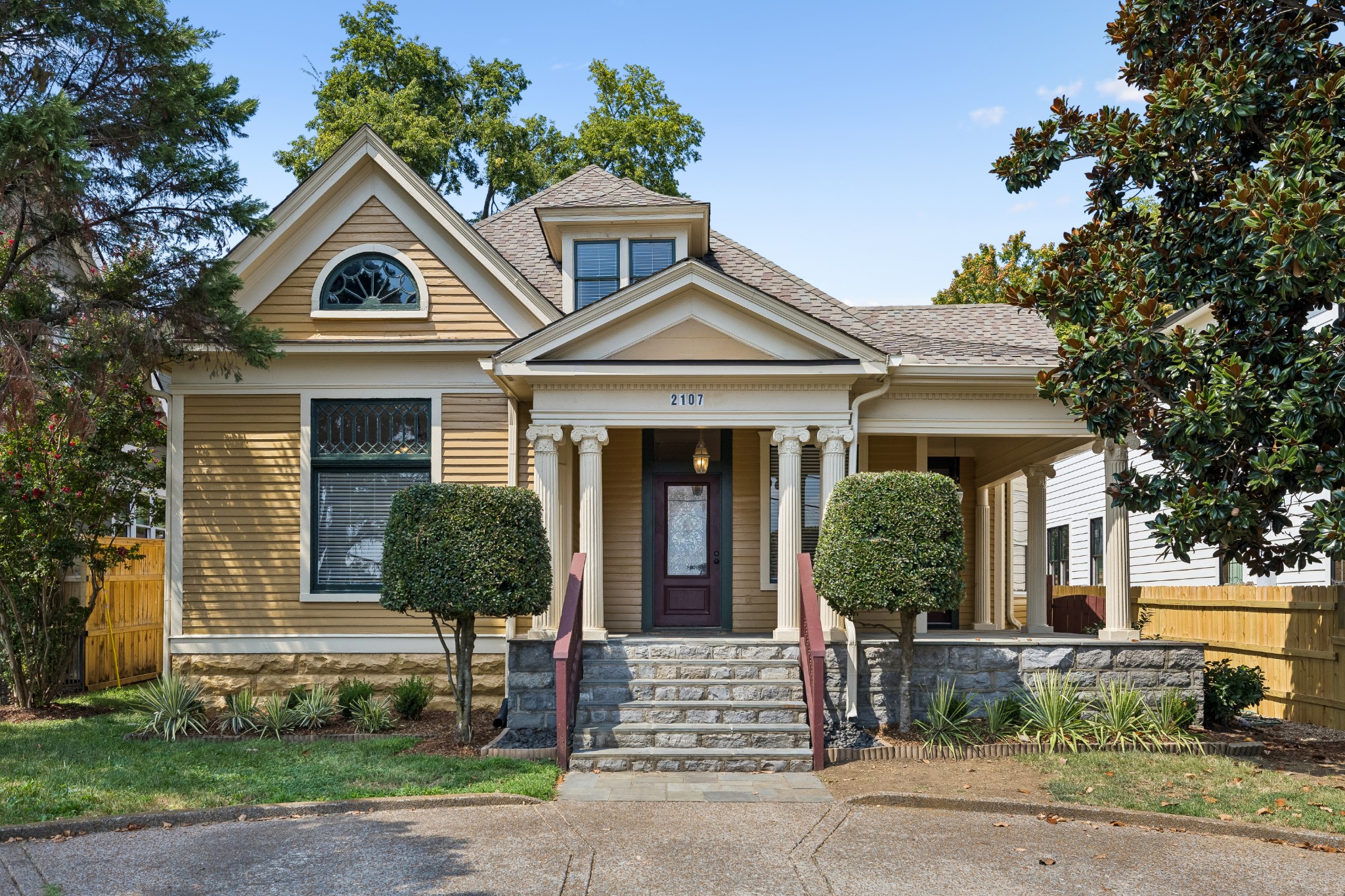 Tan two-story house with a columned porch, stone steps, and clipped shrubs flanking the entrance.