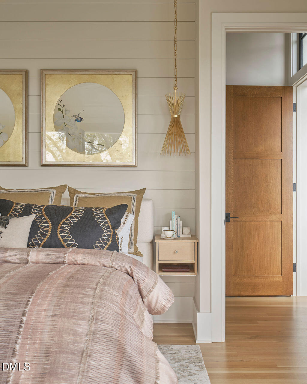 Staged bedroom with a pink quilt, patterned pillows, paneled wall, and wooden door.