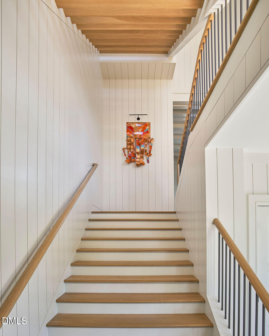 White-paneled staircase with wide wood treads, black balusters, wood ceiling beams, and colorful artwork.