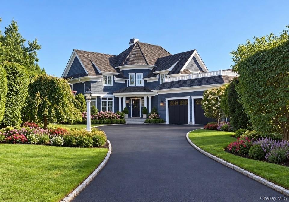 Blue-gray shingle-style home with a curved driveway, mature trees, and colorful front garden beds.