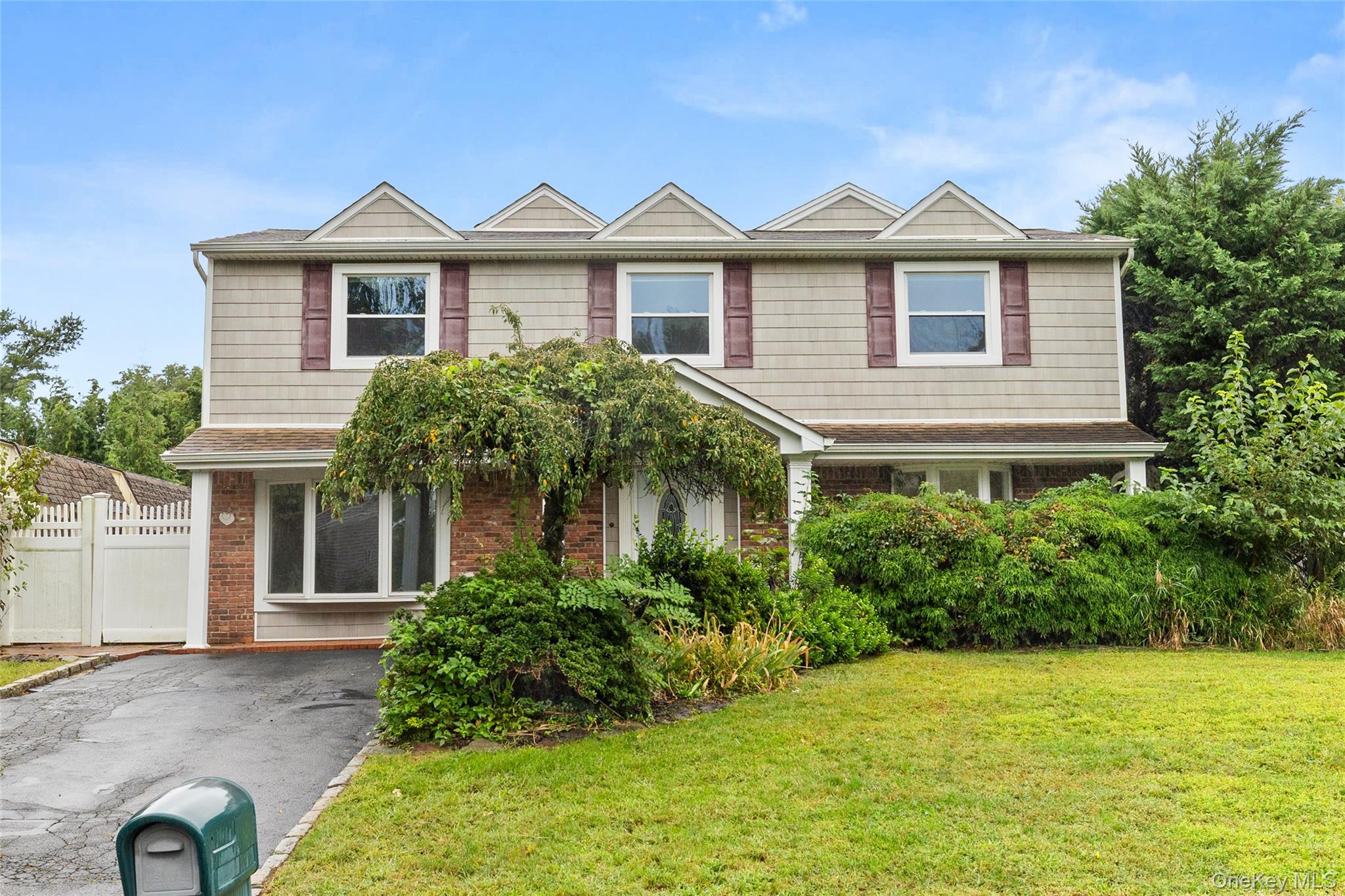 Two-story beige and brick building with a gabled roof, dense landscaping, and a driveway.