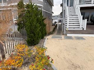 Beachfront homes flank a sandy path toward the shore, bordered by an evergreen shrub and autumn flowers.