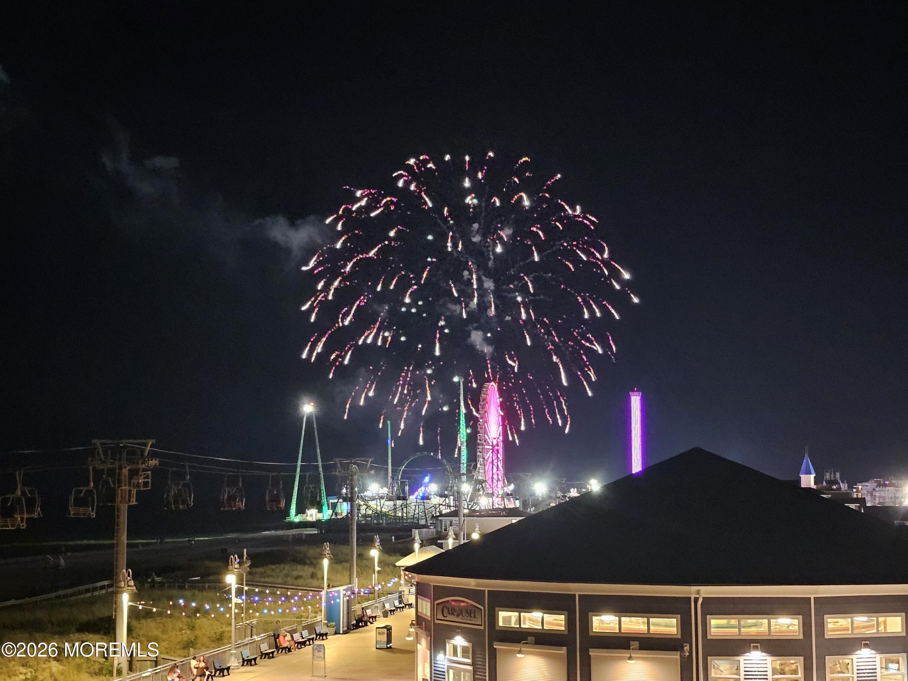 Fireworks burst above a boardwalk amusement area, with illuminated rides and the Carousel building below.