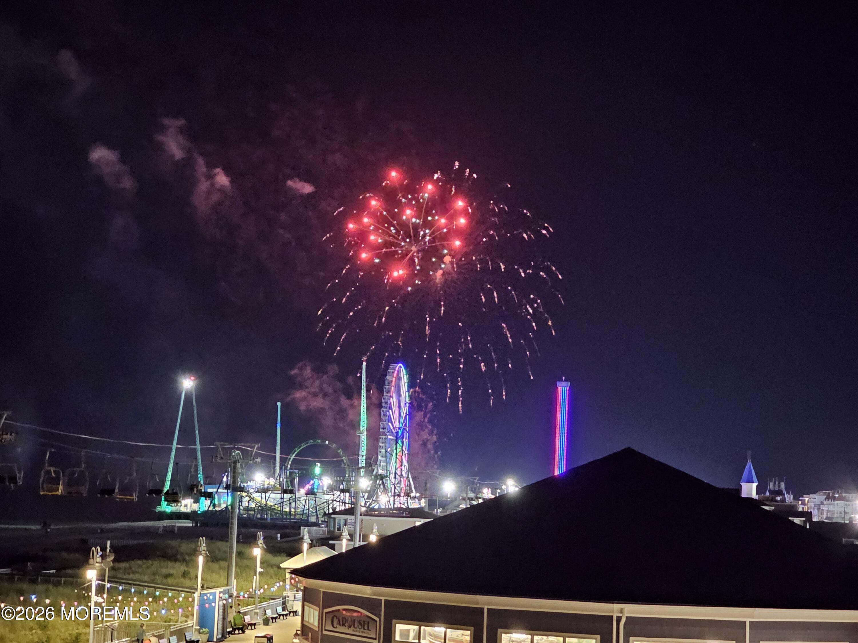 Fireworks burst above a seaside amusement park, with a Ferris wheel and illuminated rides below.