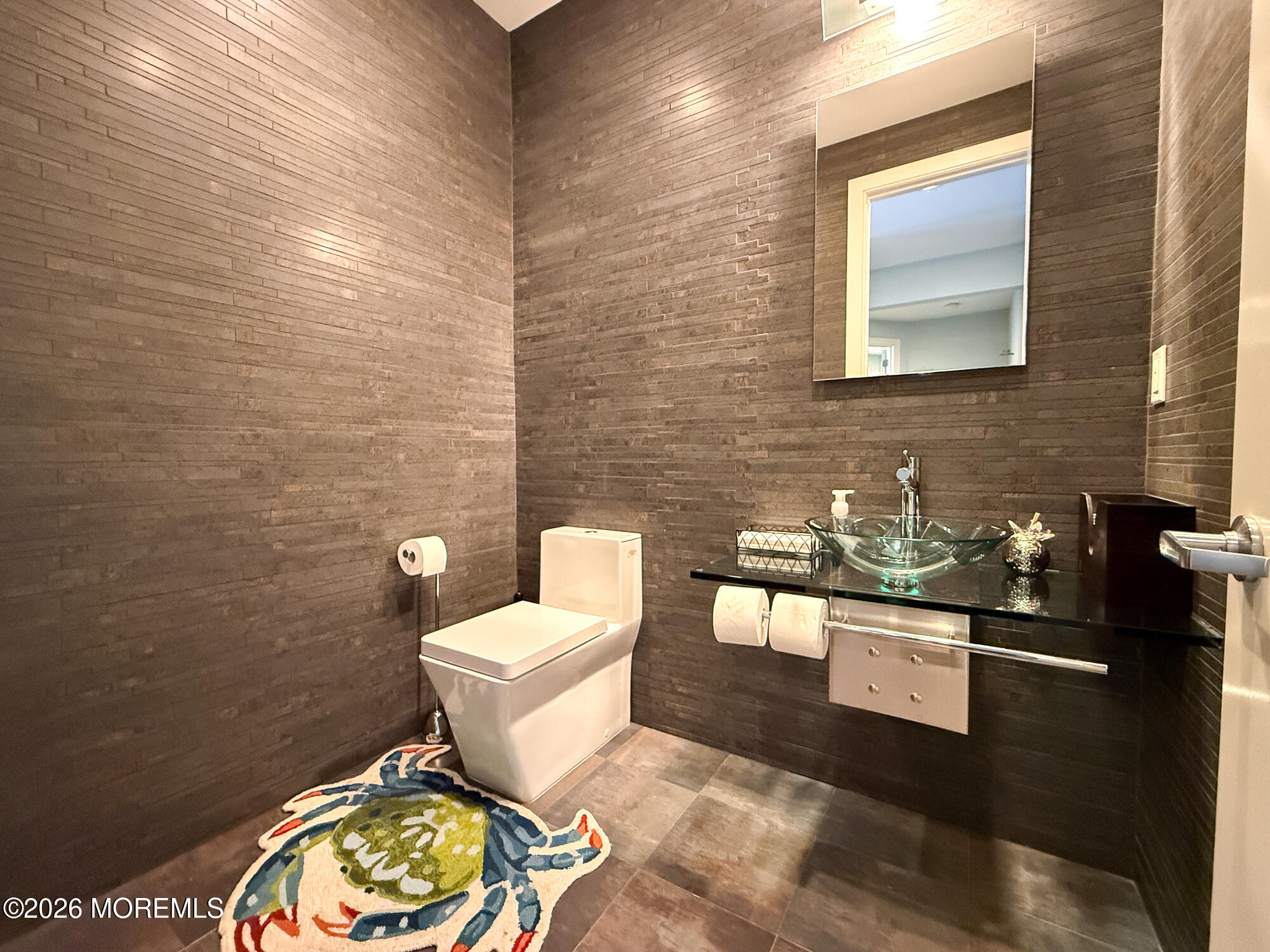 Bathroom with dark stone-tile walls, glass vessel sink, white toilet, and colorful crab-shaped rug.