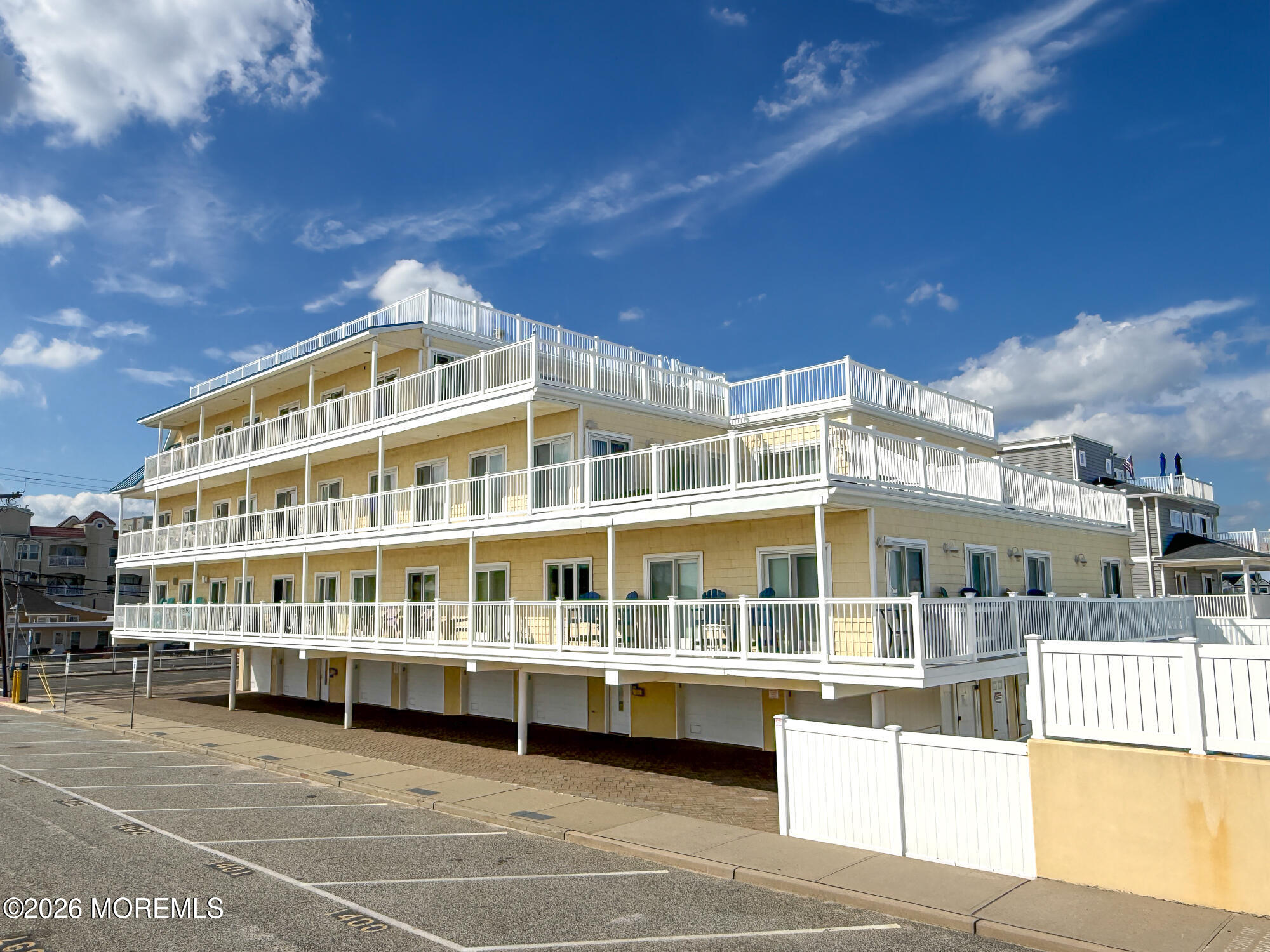 Beige multi-story coastal building with white balconies, rooftop terraces, and parking spaces in front.