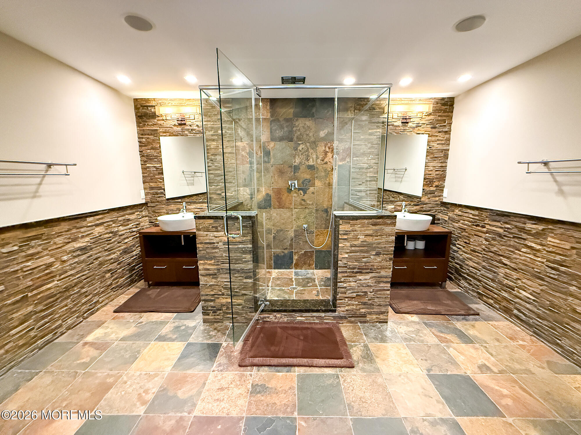 Bathroom with a glass walk-in shower and matching vessel sinks against stone tile.