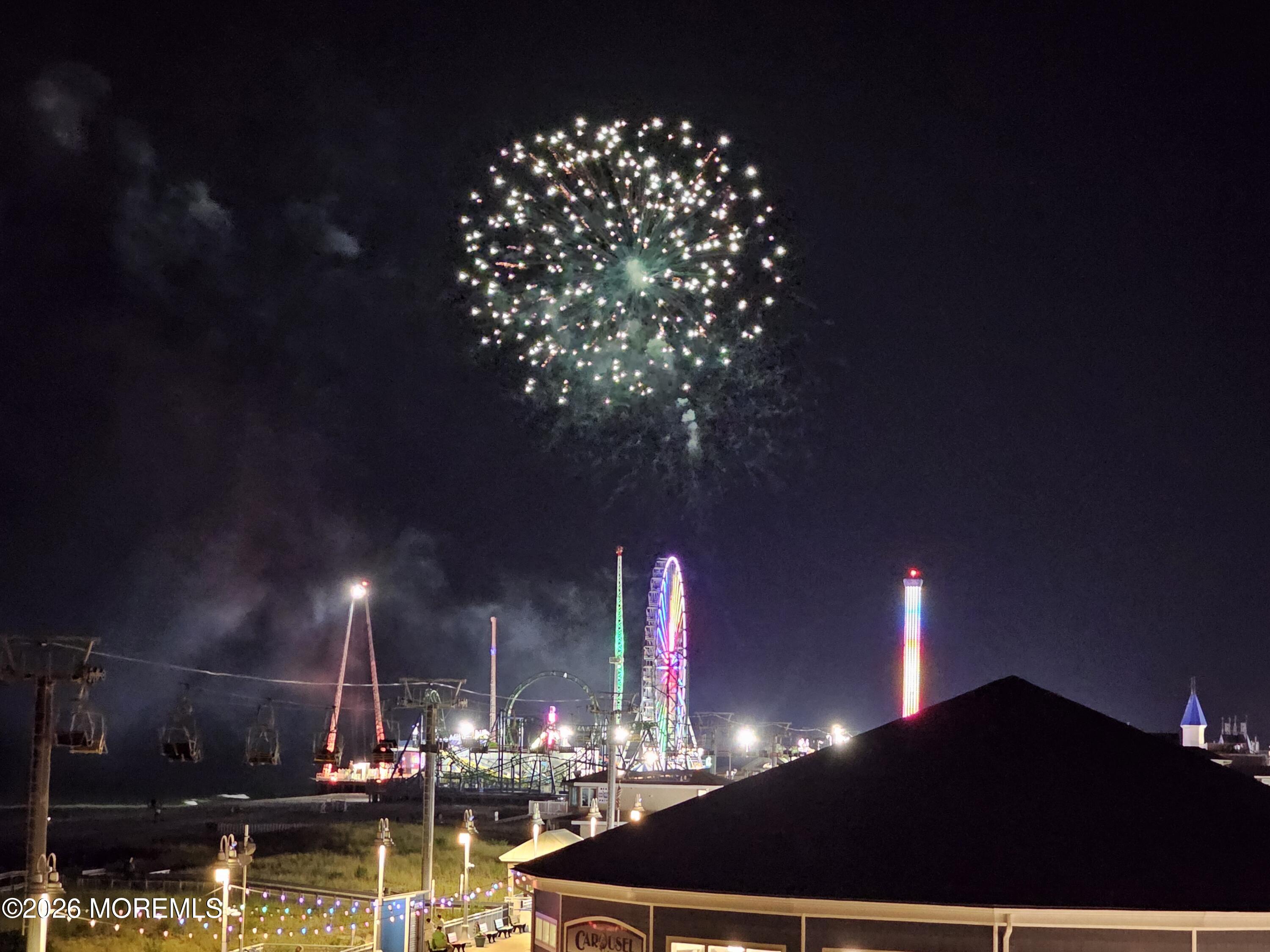 Fireworks above Seaside Heights boardwalk rides, with a lit Ferris wheel and carousel roof.