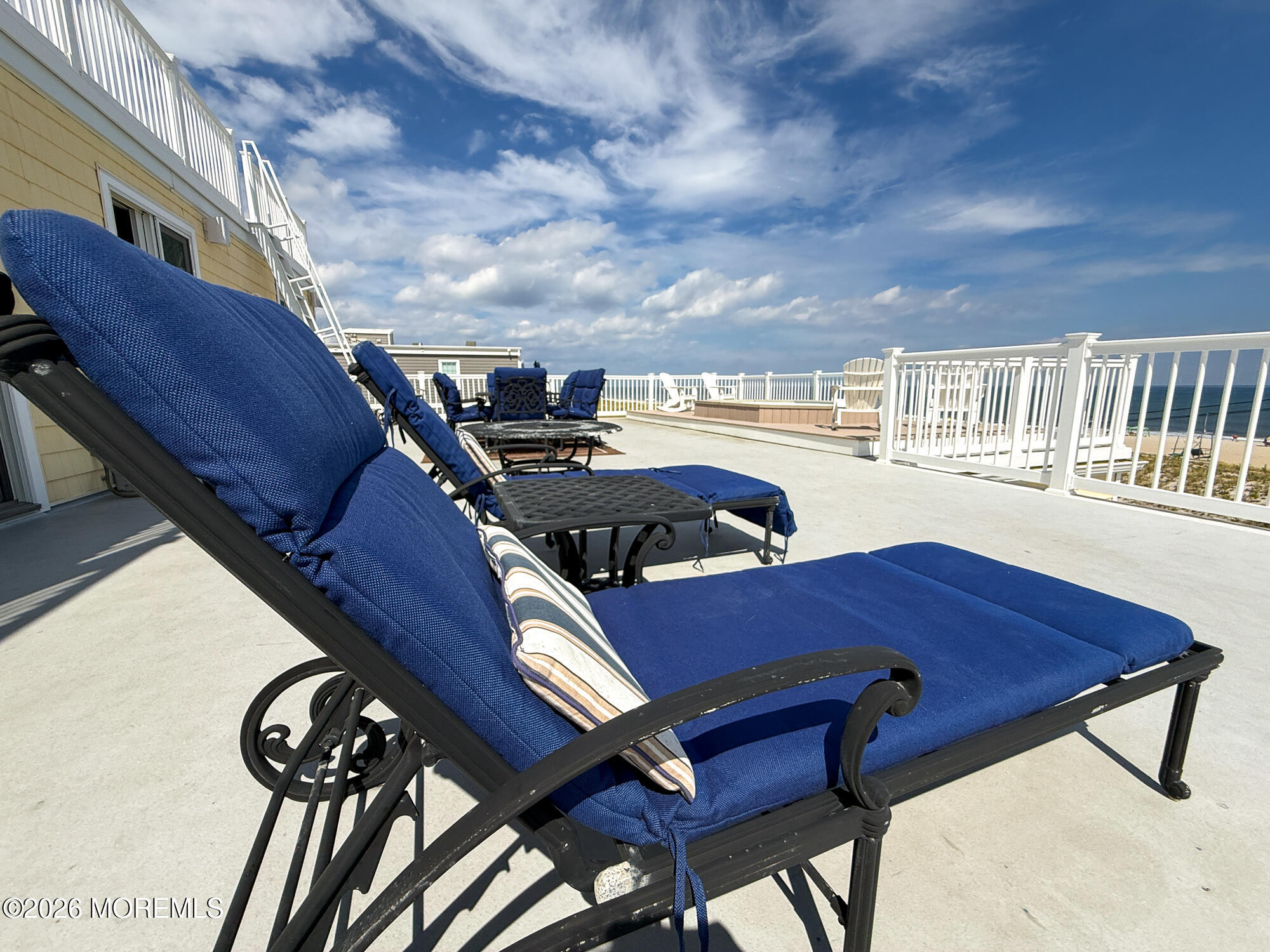 Oceanfront terrace with blue-cushioned lounge chairs, white railings, and a view of the beach and ocean.