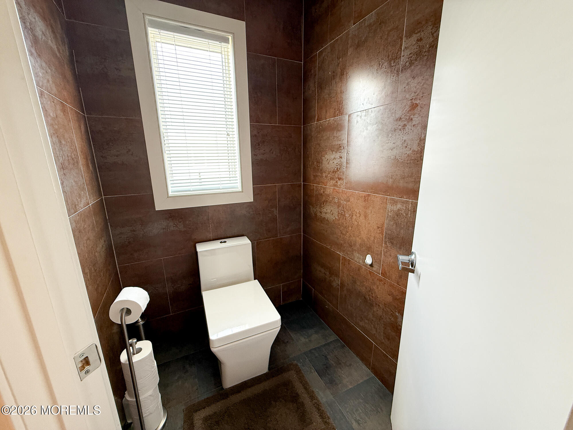 Bathroom with a white toilet, dark brown tiled walls, and a high window with blinds.