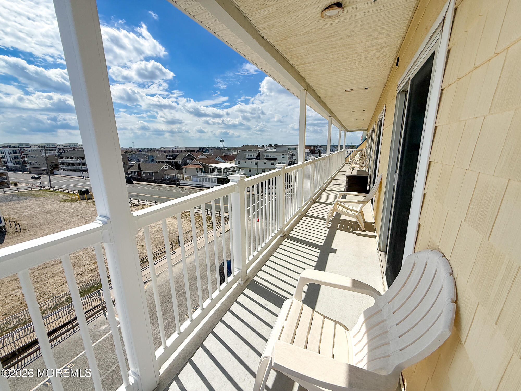 Furnished balcony overlooking neighborhood rooftops, with white chairs and a long striped railing shadow.