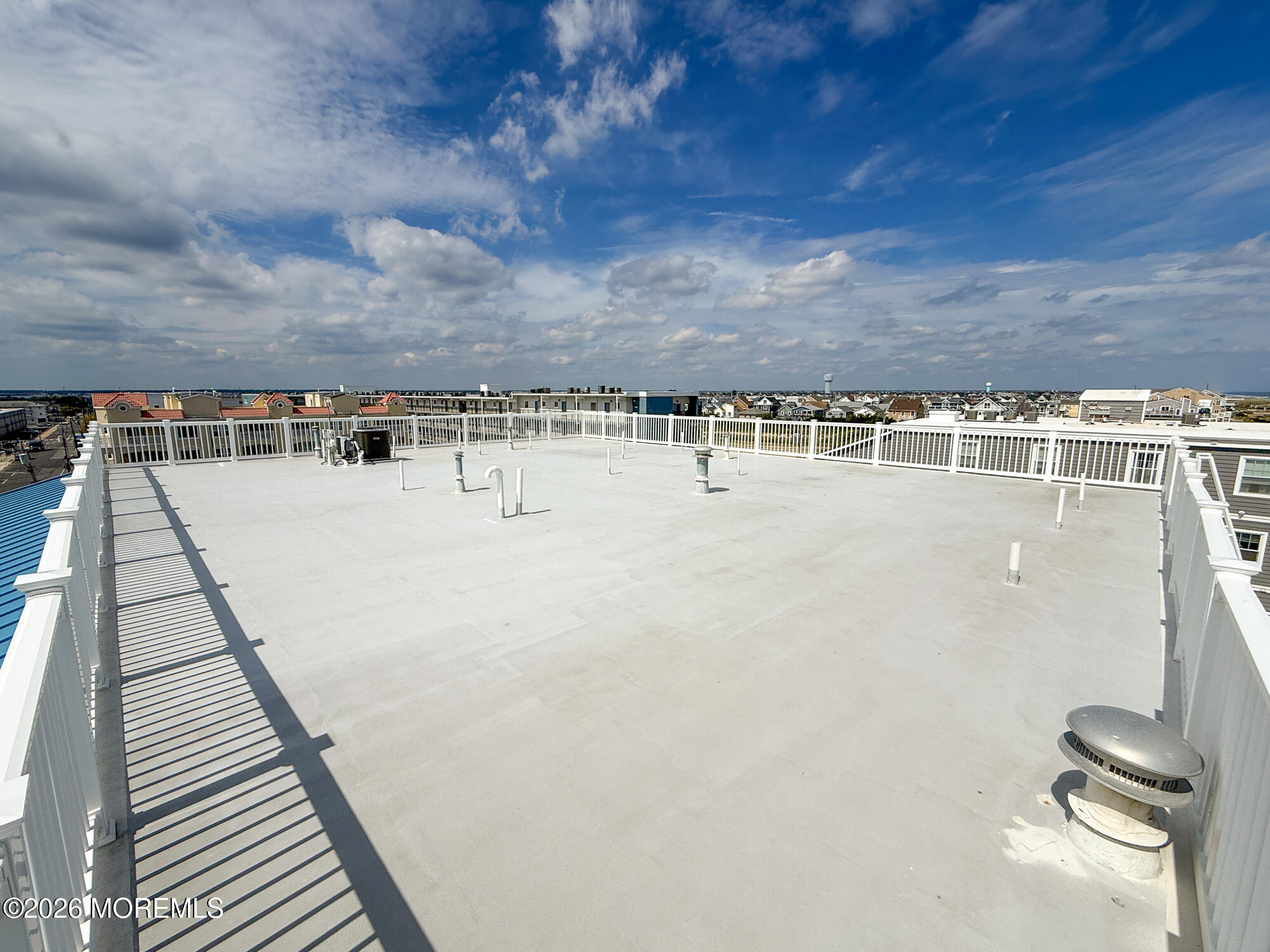Expansive flat rooftop terrace with white railings and ventilation fixtures, overlooking nearby homes.