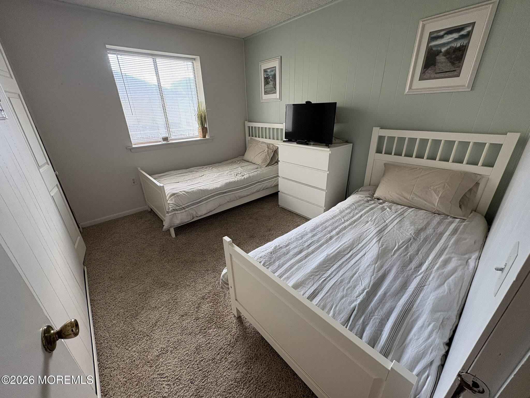 Bedroom with two white beds, a dresser and television, and a window with blinds.