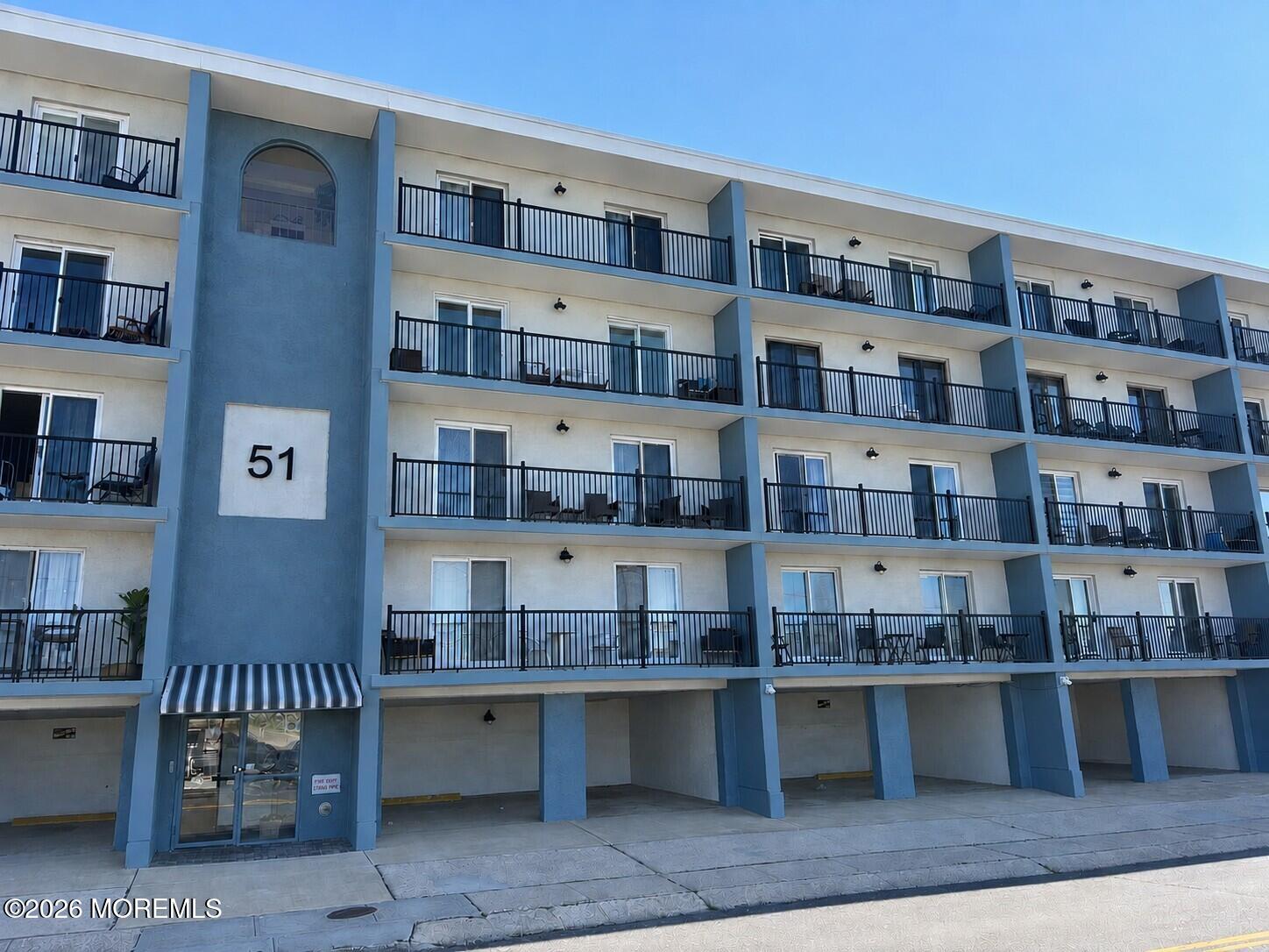 Multi-story condominium building with blue-and-white facade, rows of balconies, and covered ground-level parking.
