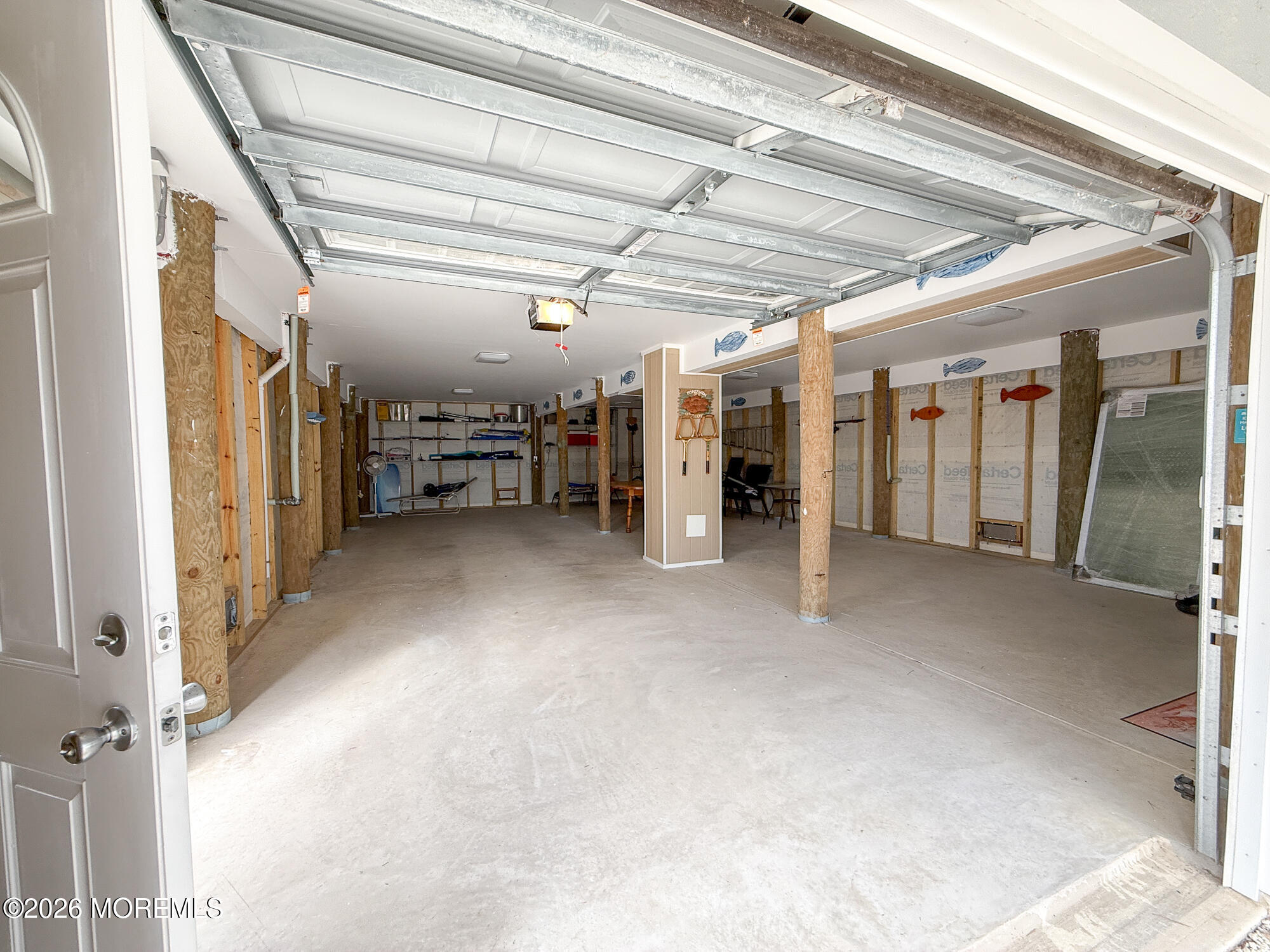 Large garage with an open overhead door, concrete floor, exposed framing, and storage shelves.
