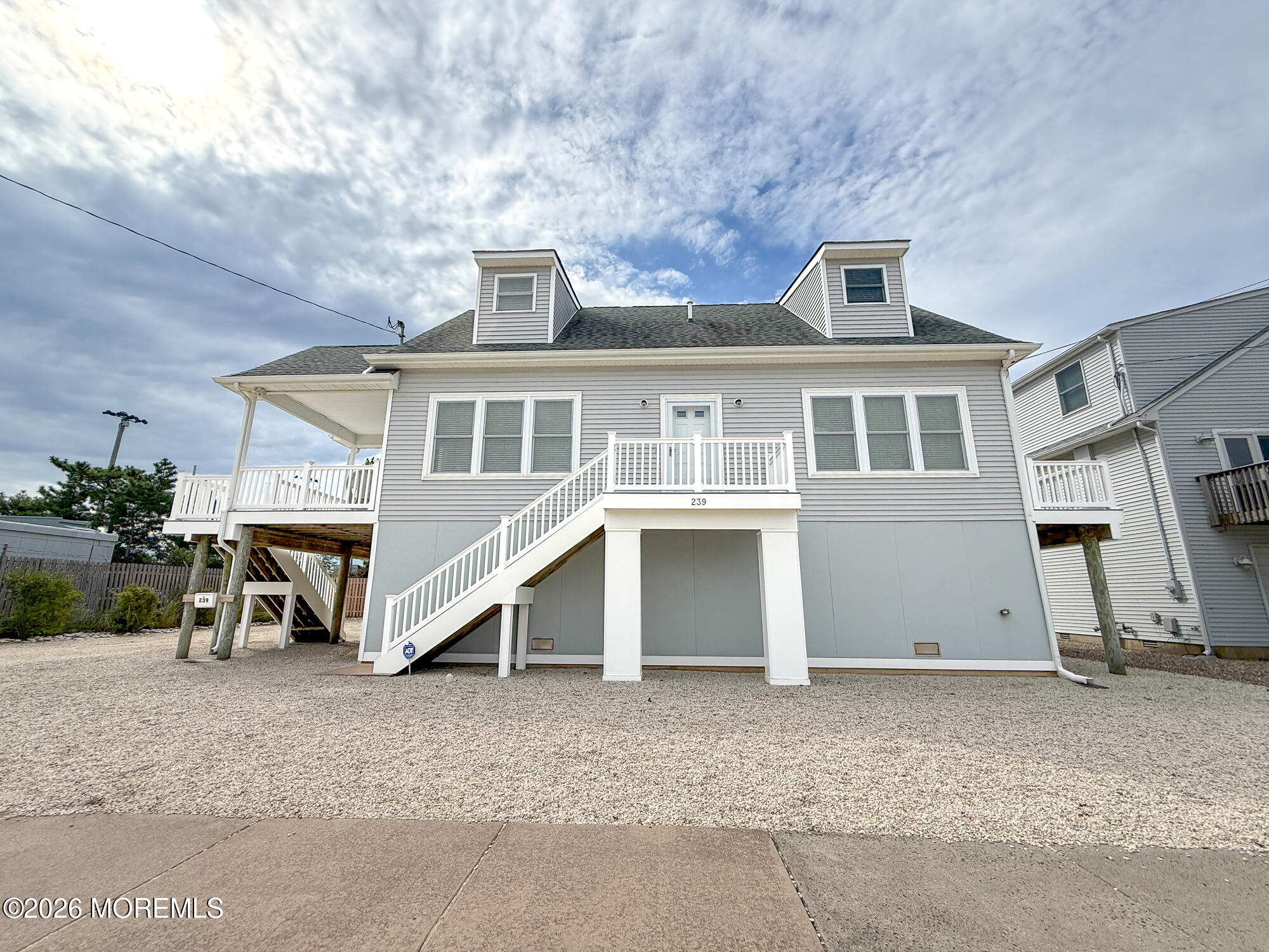 Raised gray coastal home with white stairs, porch railings, and gravel yard.