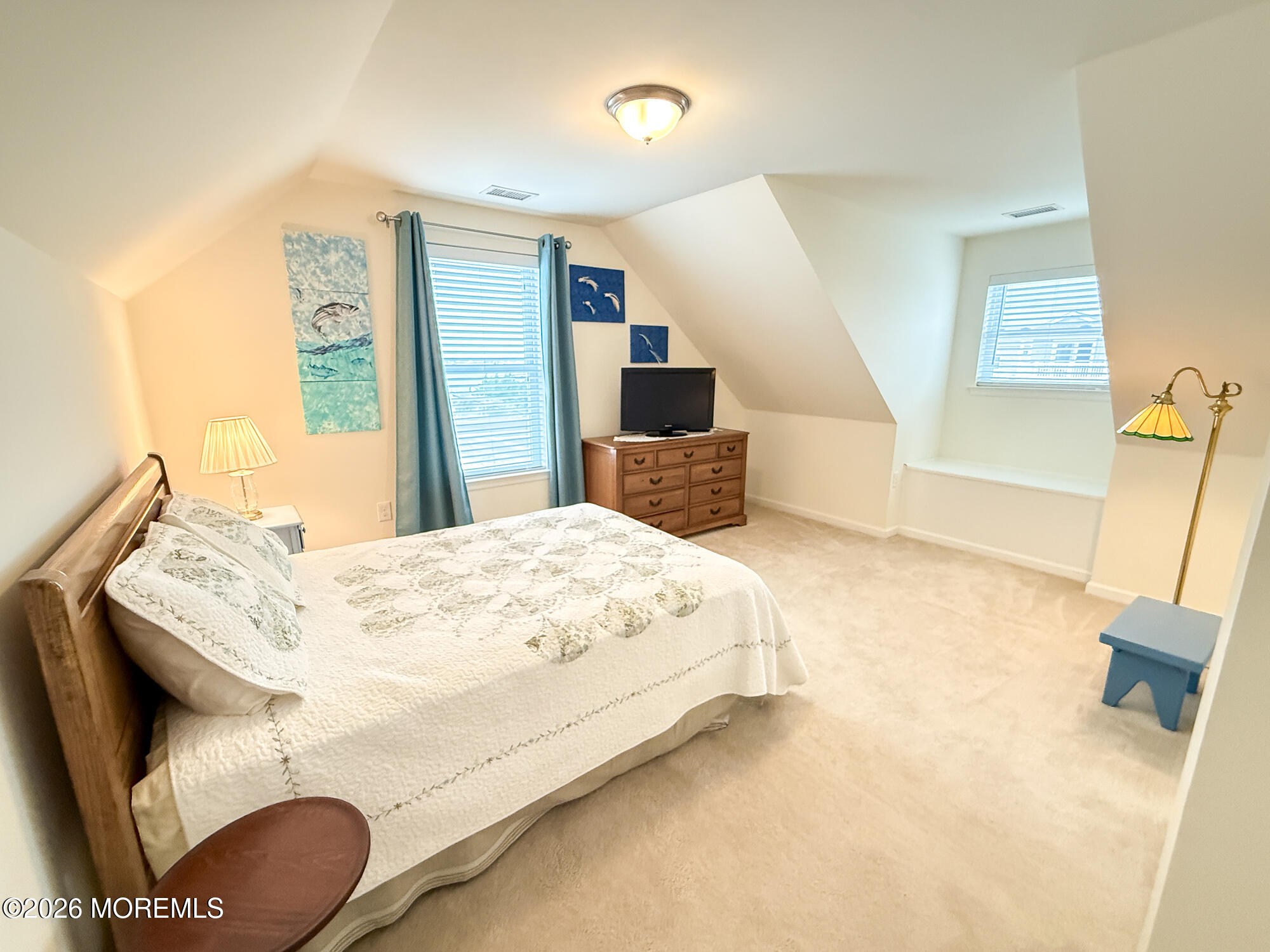 Furnished bedroom with a large bed, sloped ceilings, and a wooden dresser beneath coastal artwork.