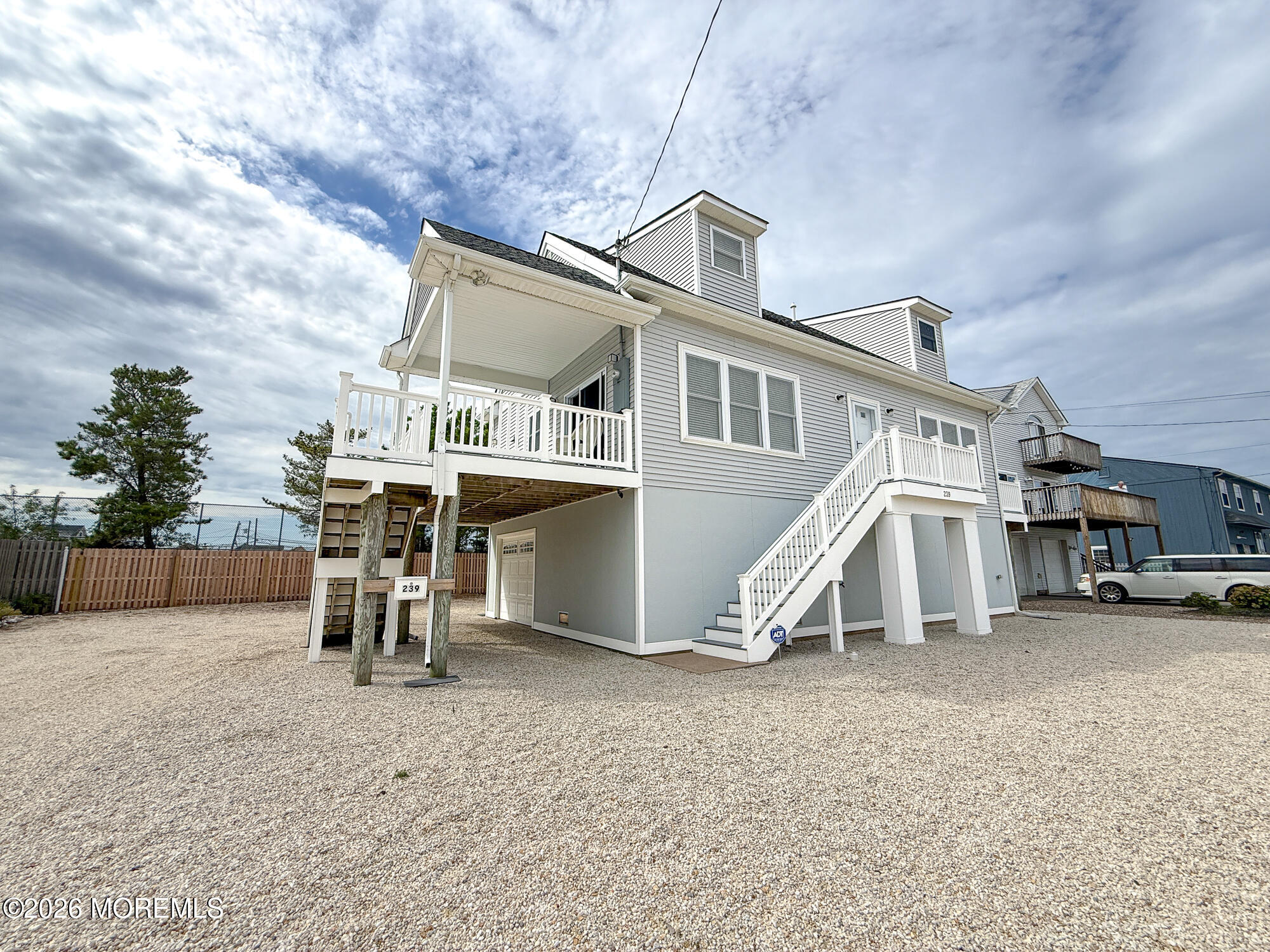 Raised gray house with white trim, covered porch, exterior stairs, and a broad gravel lot.