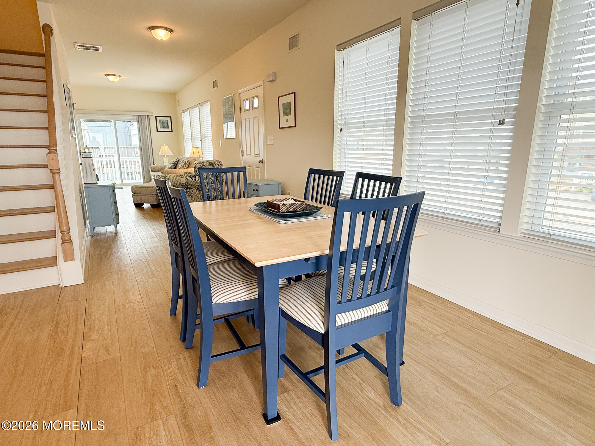 Dining area with a light wood table and blue chairs beside large windows and a staircase.
