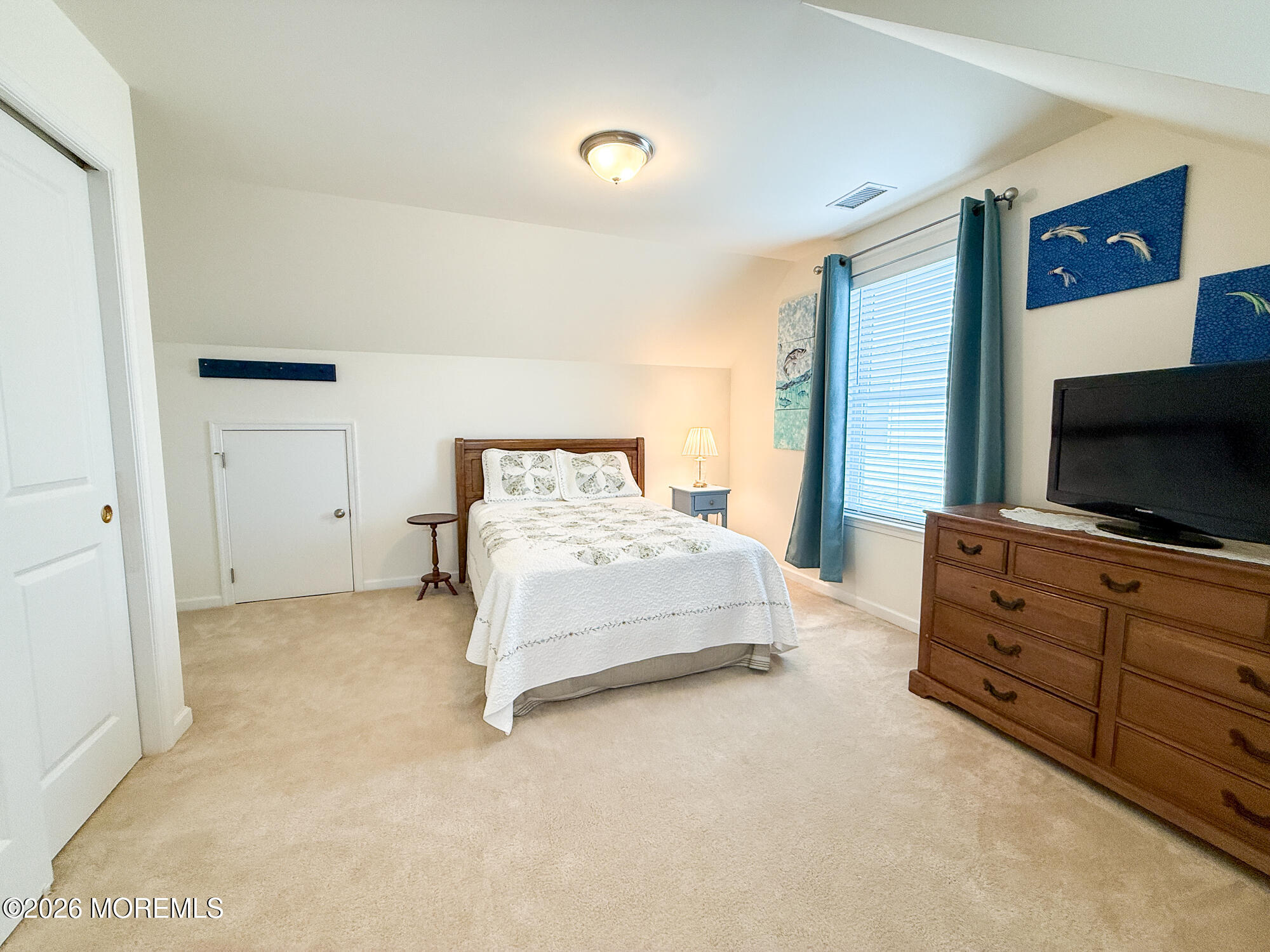 Furnished bedroom with a white bed, beige carpet, teal curtains, and a wooden dresser with television.