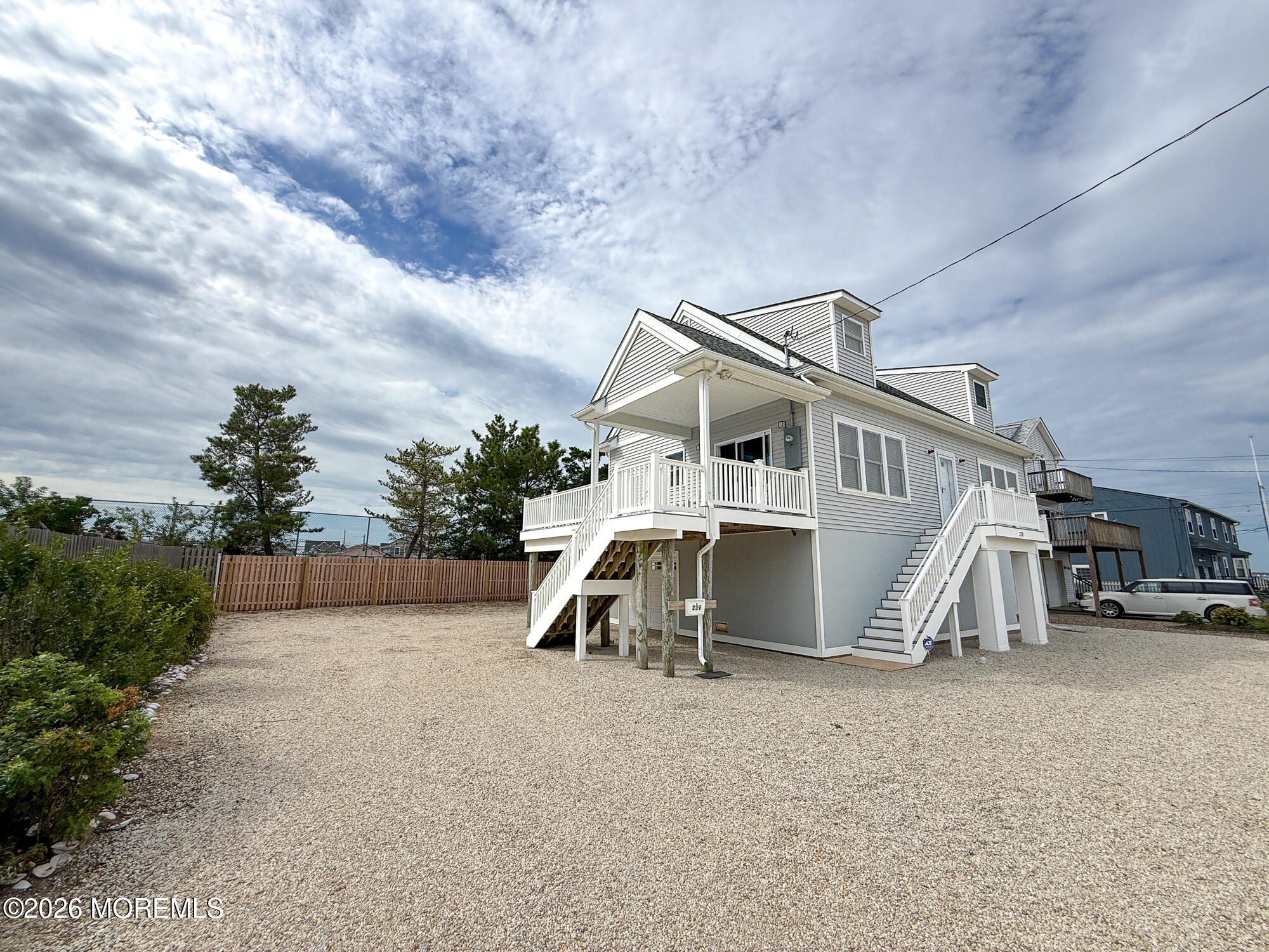 Raised gray coastal house with white porches and stairs above a broad gravel yard.