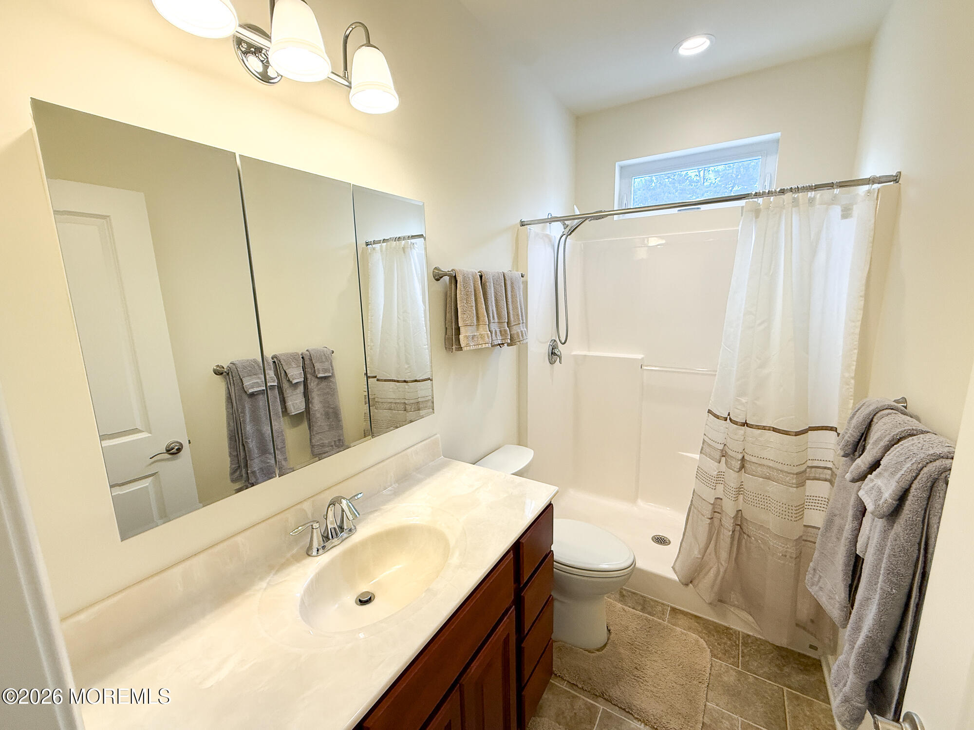 Bathroom with a long mirrored vanity, toilet, and curtained shower beneath a window.