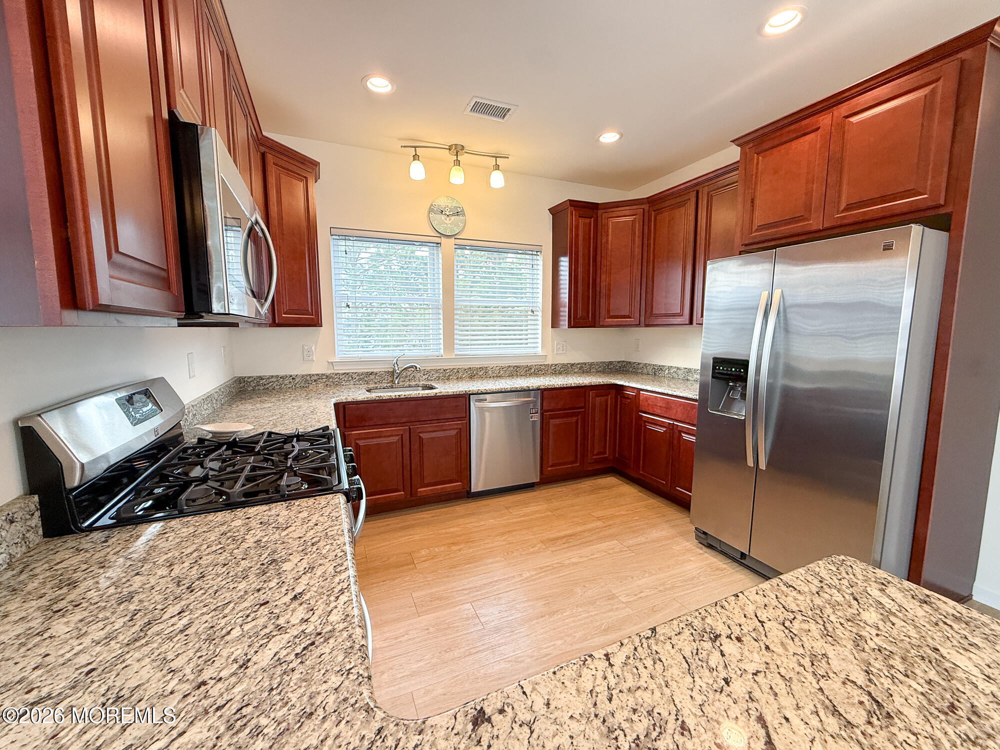 Kitchen with reddish wood cabinets, granite counters, stainless steel appliances, and windows above the sink.