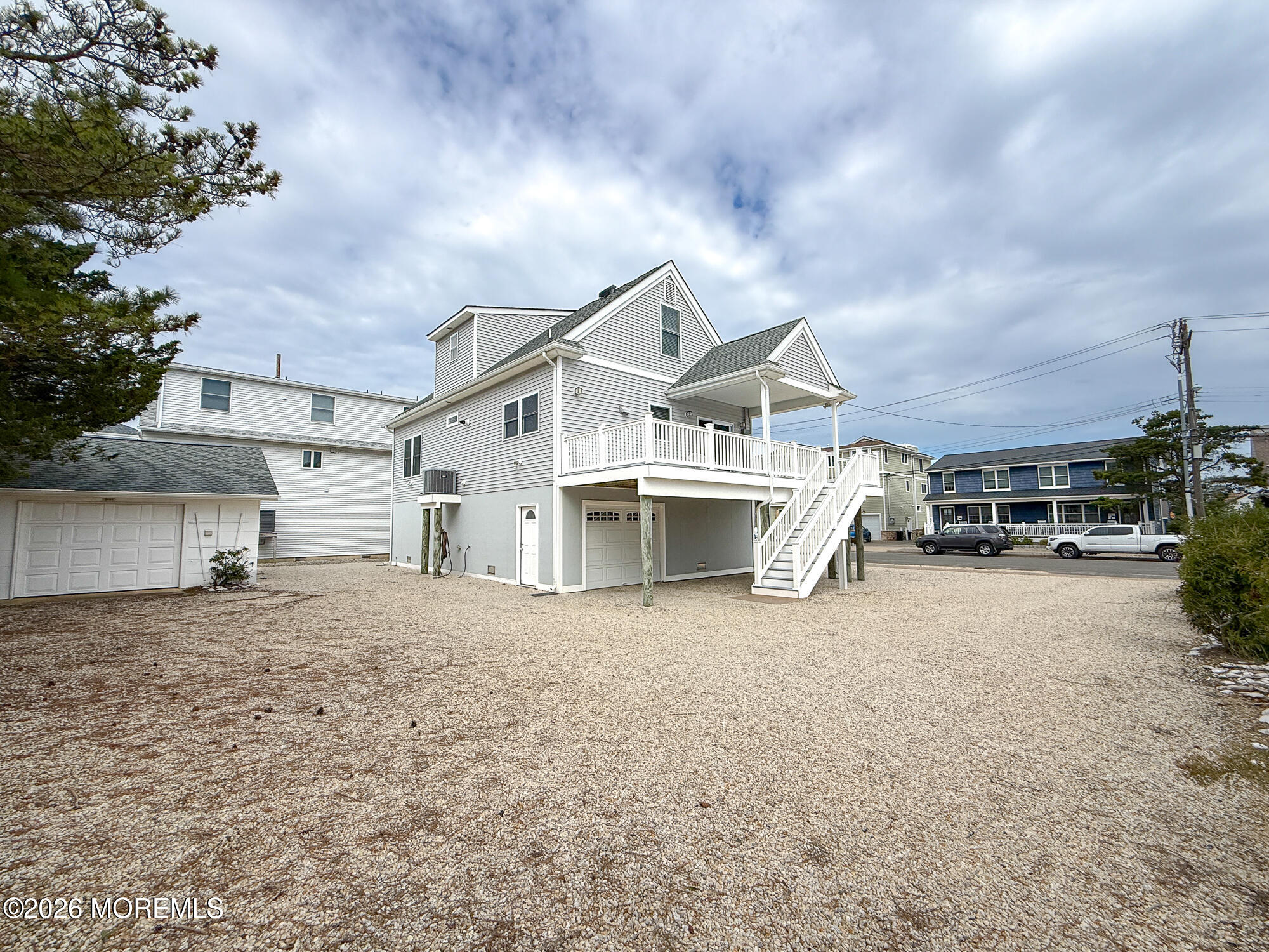 White multi-story house with a raised deck, exterior staircase, garage, and broad gravel yard.