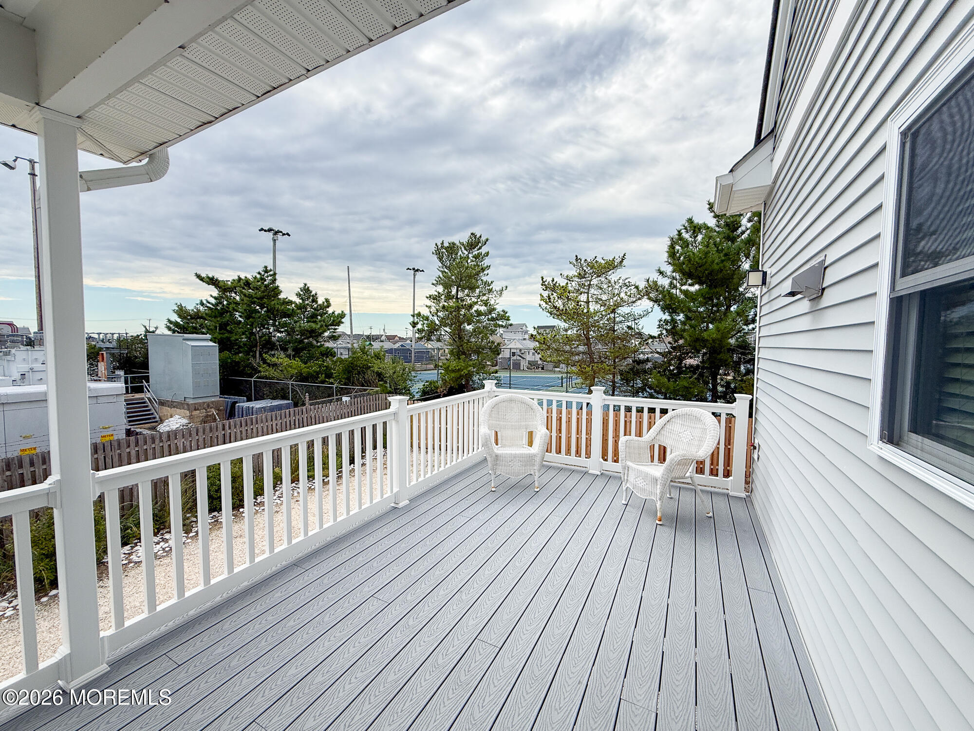 Elevated deck with gray composite flooring, white railings, and two wicker chairs overlooking tennis courts.