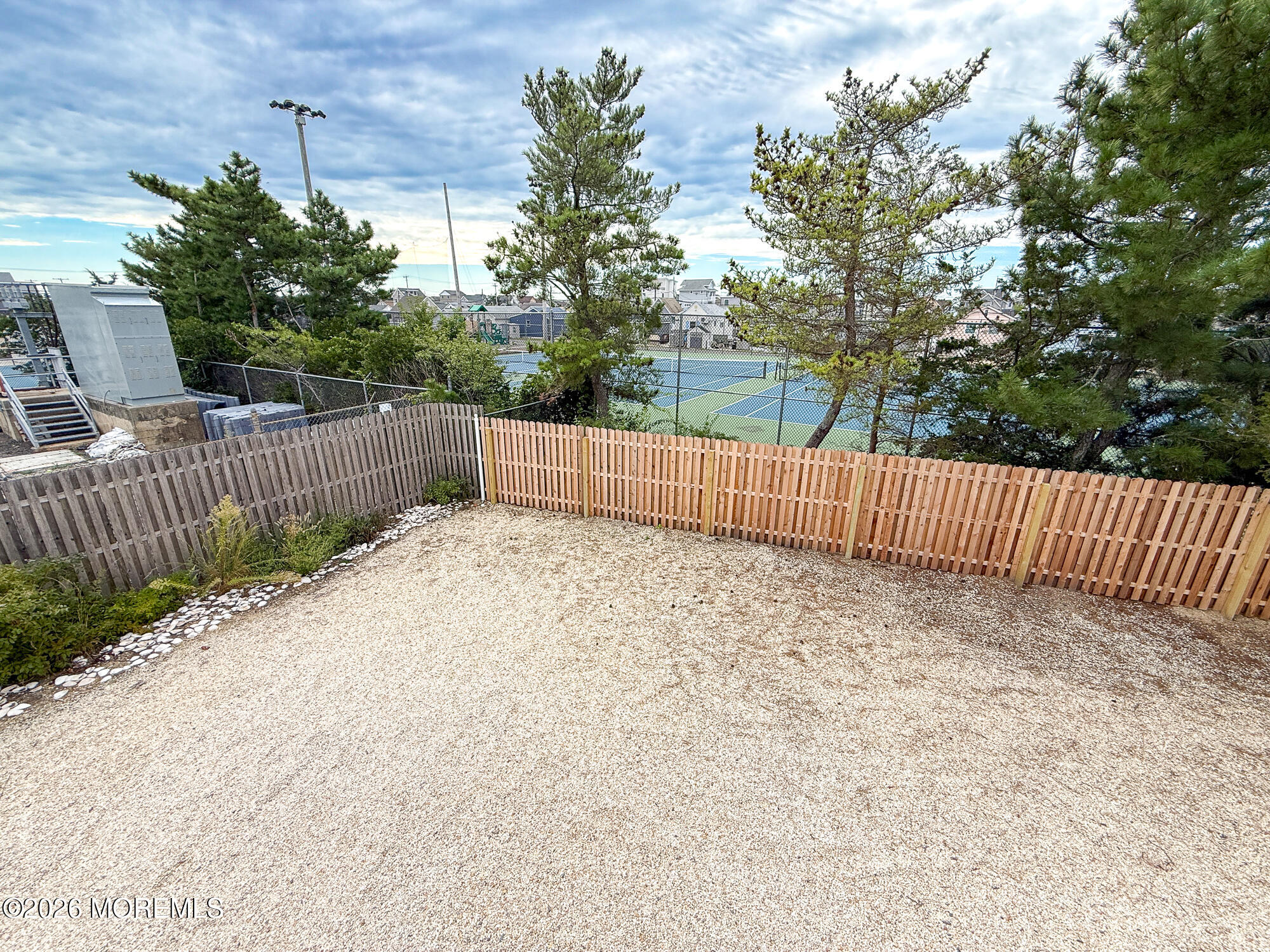 Gravel backyard with a wooden fence, mature trees, and tennis courts beyond.
